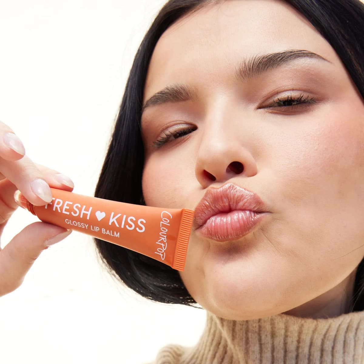 Dark-haired model puckering glossy nude-amber lips while holding an orange ColourPop Fresh Kiss tube to her mouth