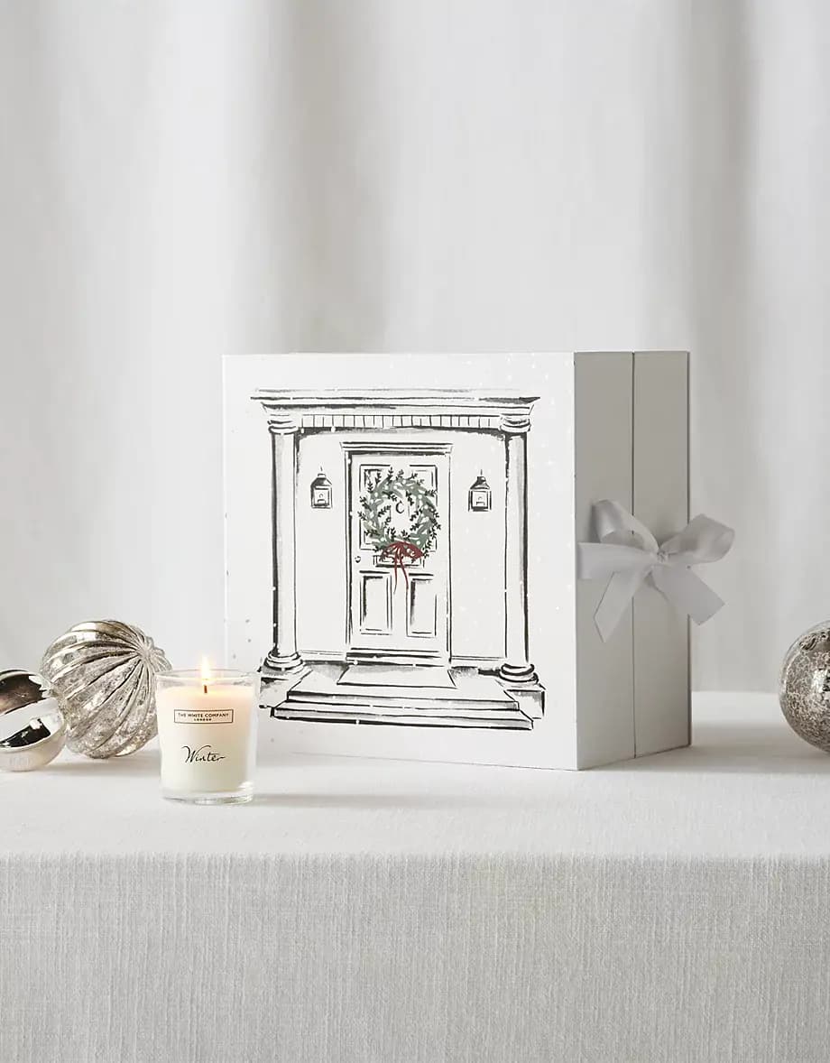 Outer box illustrated with a front door and wreath, tied with a grey ribbon, beside a lit Winter votive candle