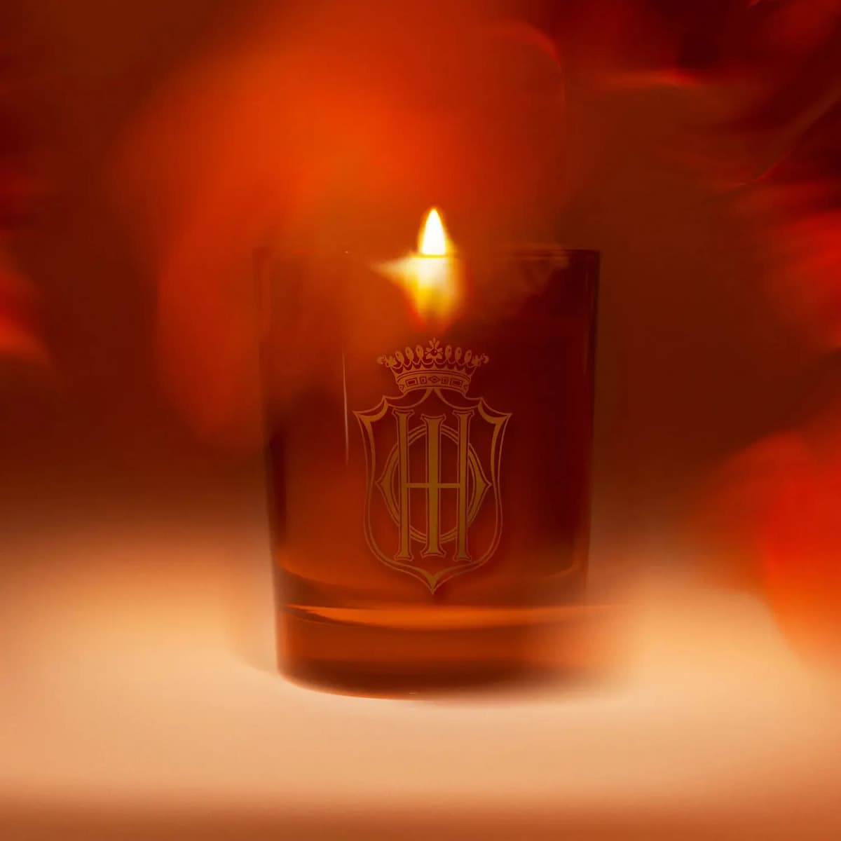 Lit candle glowing in amber glass with gold crowned monogram crest against a blurred red and orange backdrop