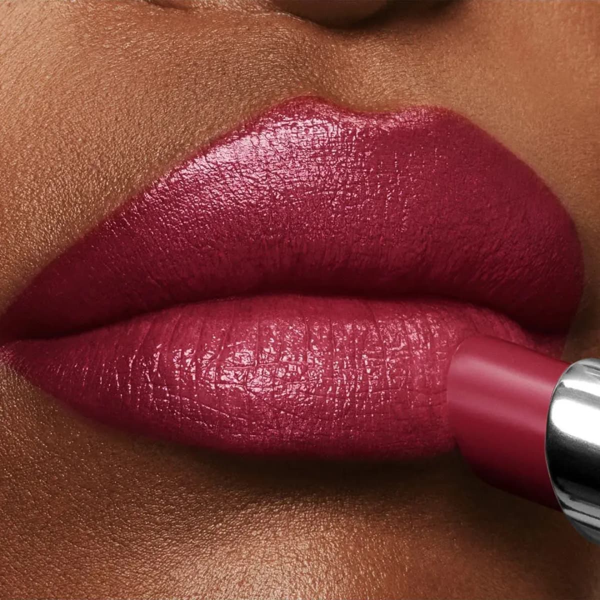 Berry-red lipstick being applied to full lips on deep skin, giving a satin shine