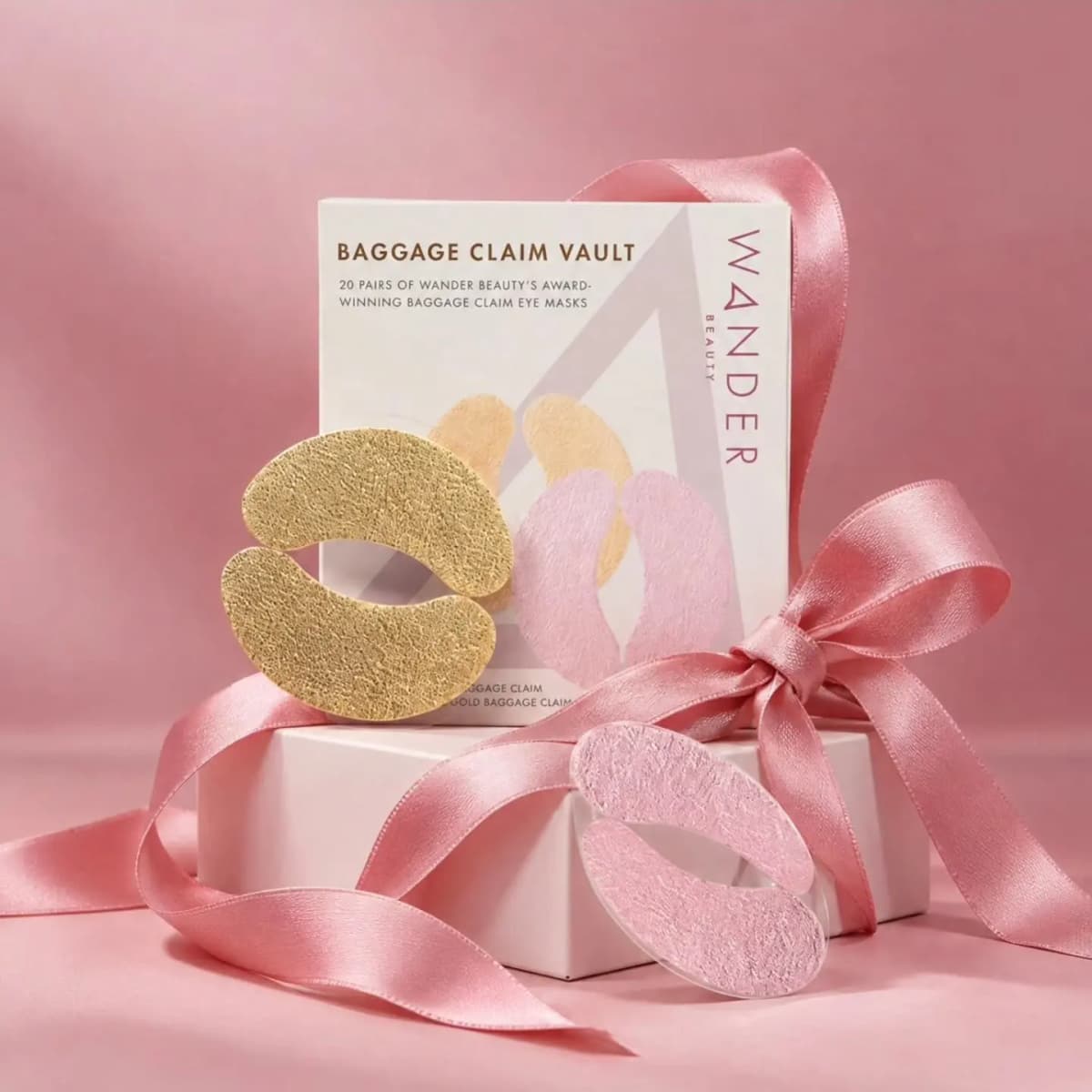 Wander Beauty Baggage Claim Eye Mask Vault box with gold and pink foil eye masks and a pink satin ribbon on a pink backdrop