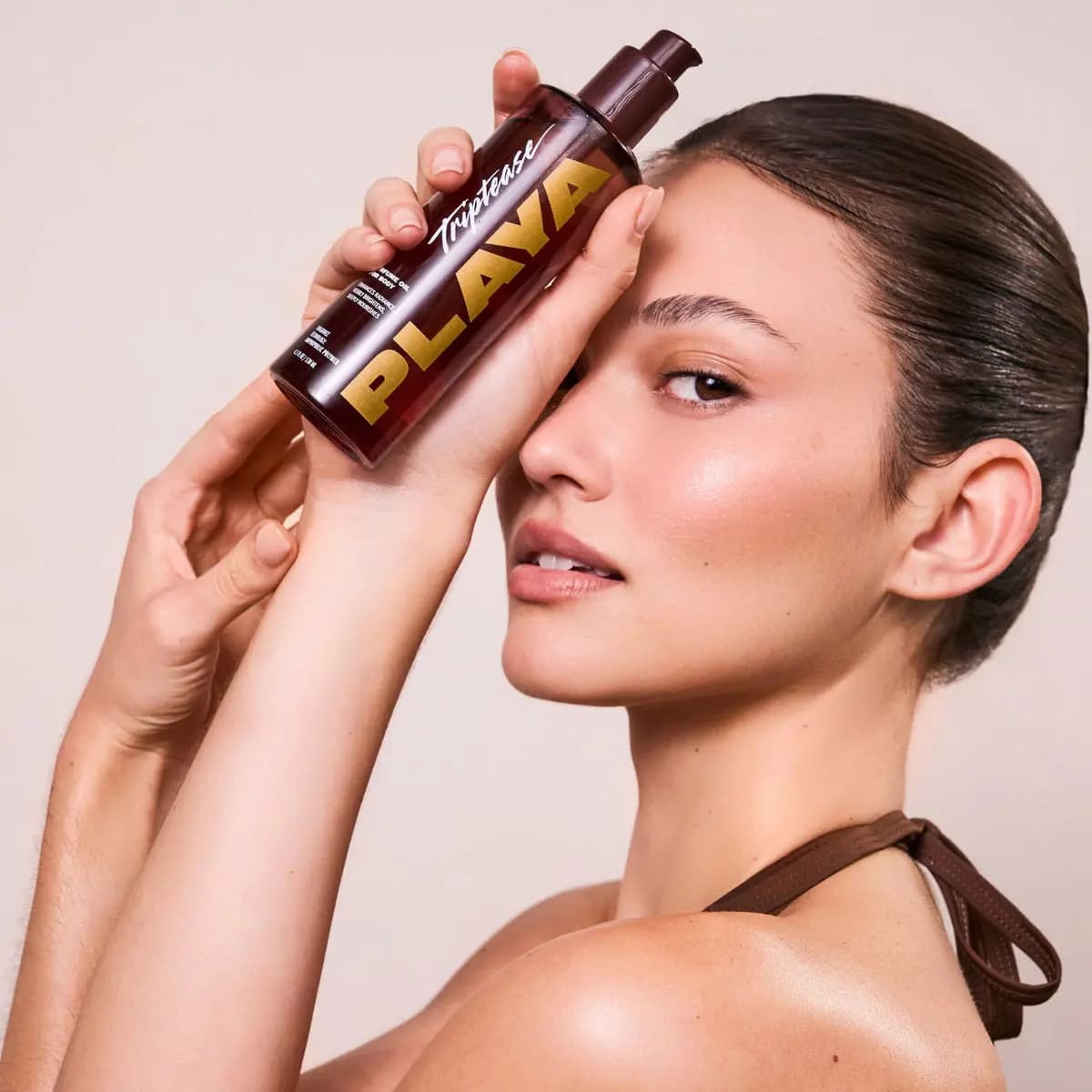Model with slicked-back hair and dewy skin holding the Triptease body oil bottle against the forehead