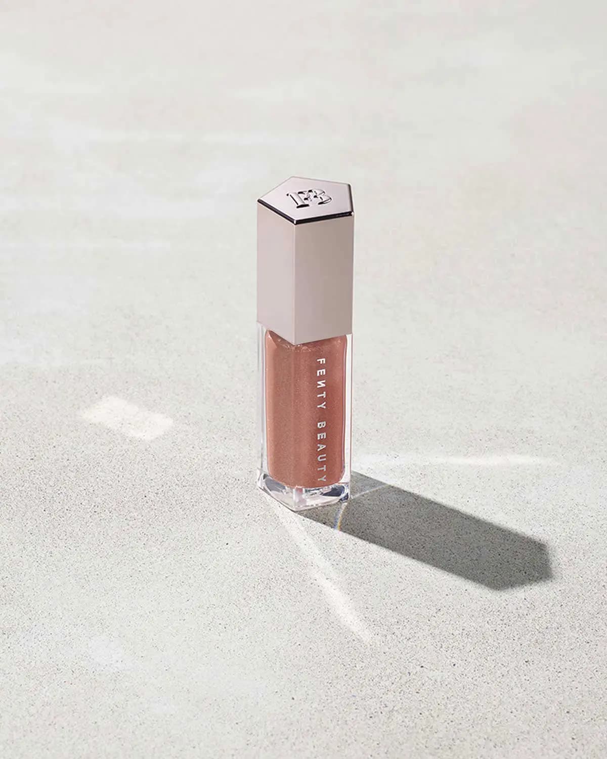 Clear lip gloss tube filled with shimmery rose-nude gloss under a pale pink hexagonal FB cap, casting a long shadow