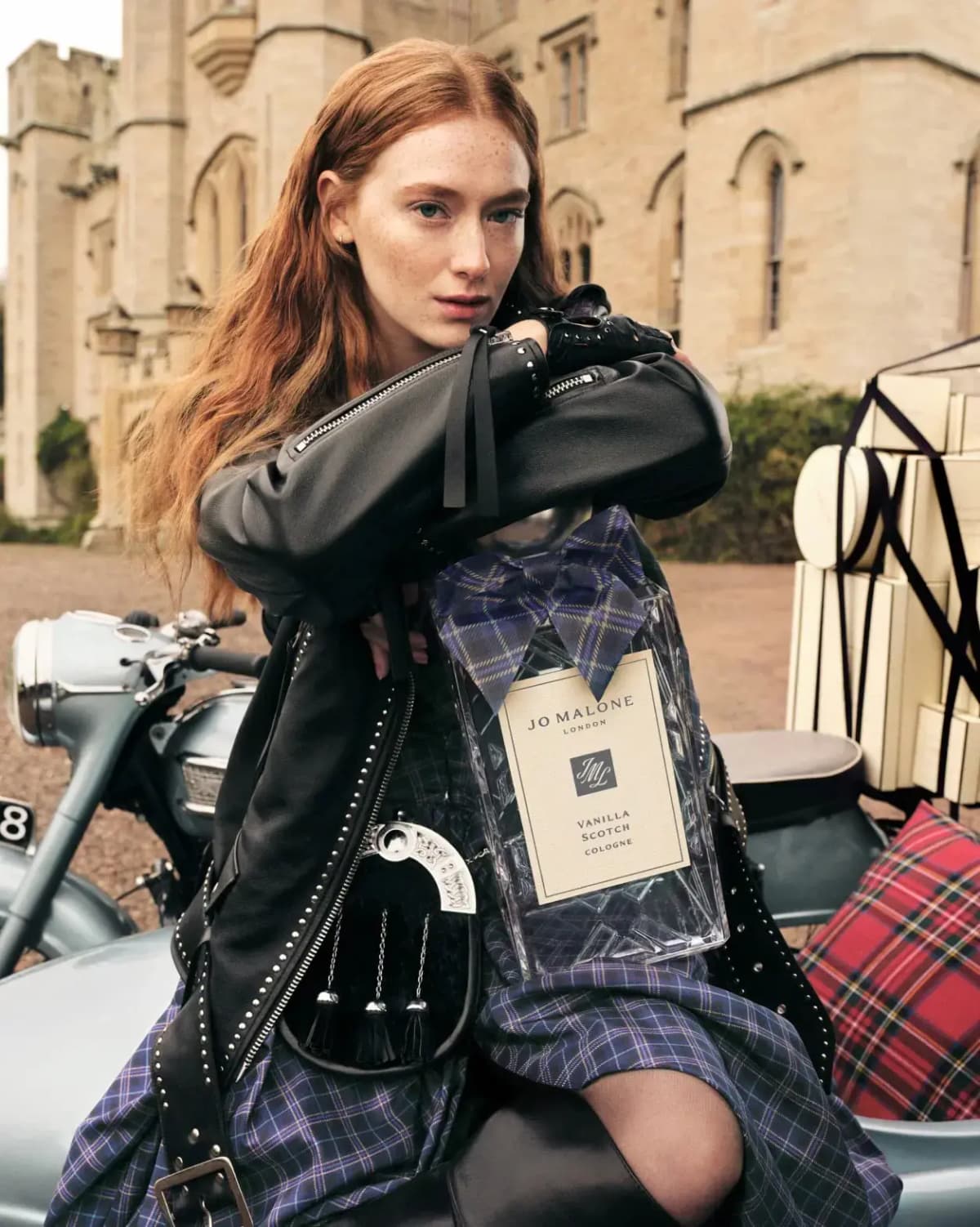 Red-haired model in leather jacket and purple tartan kilt holds an oversized cologne bottle on a motorbike outside a castle