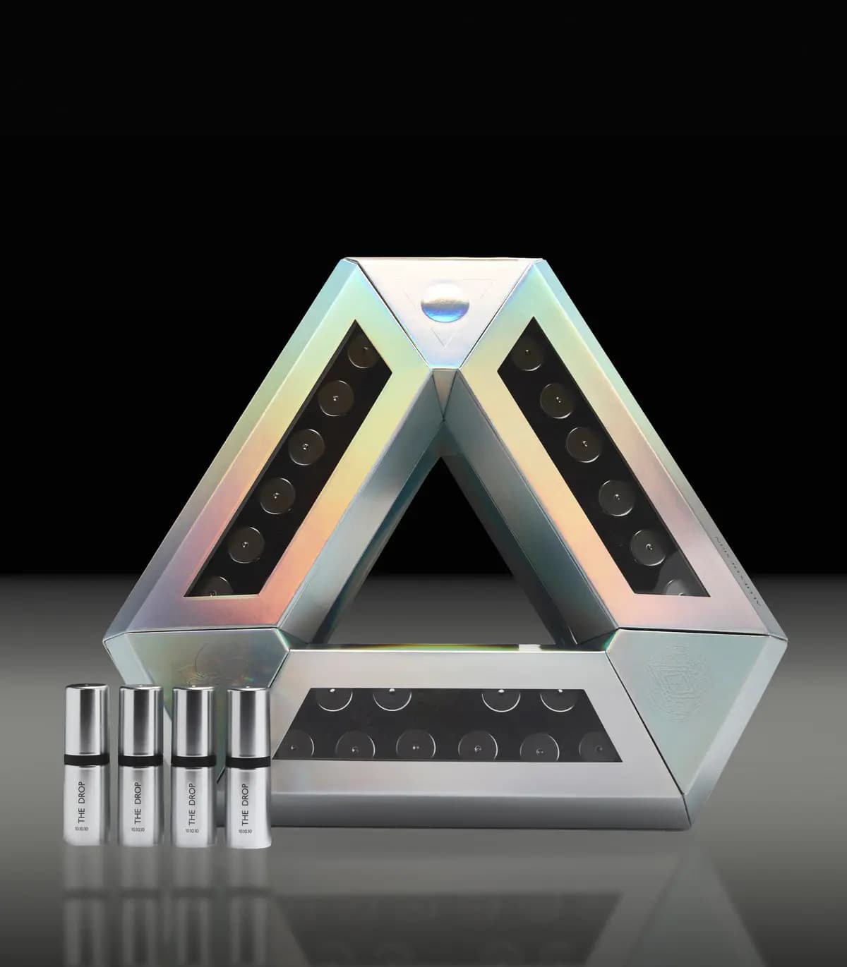 Closed holographic silver triangle case with four silver The Drop 10.10.10 ampoules on a reflective grey surface