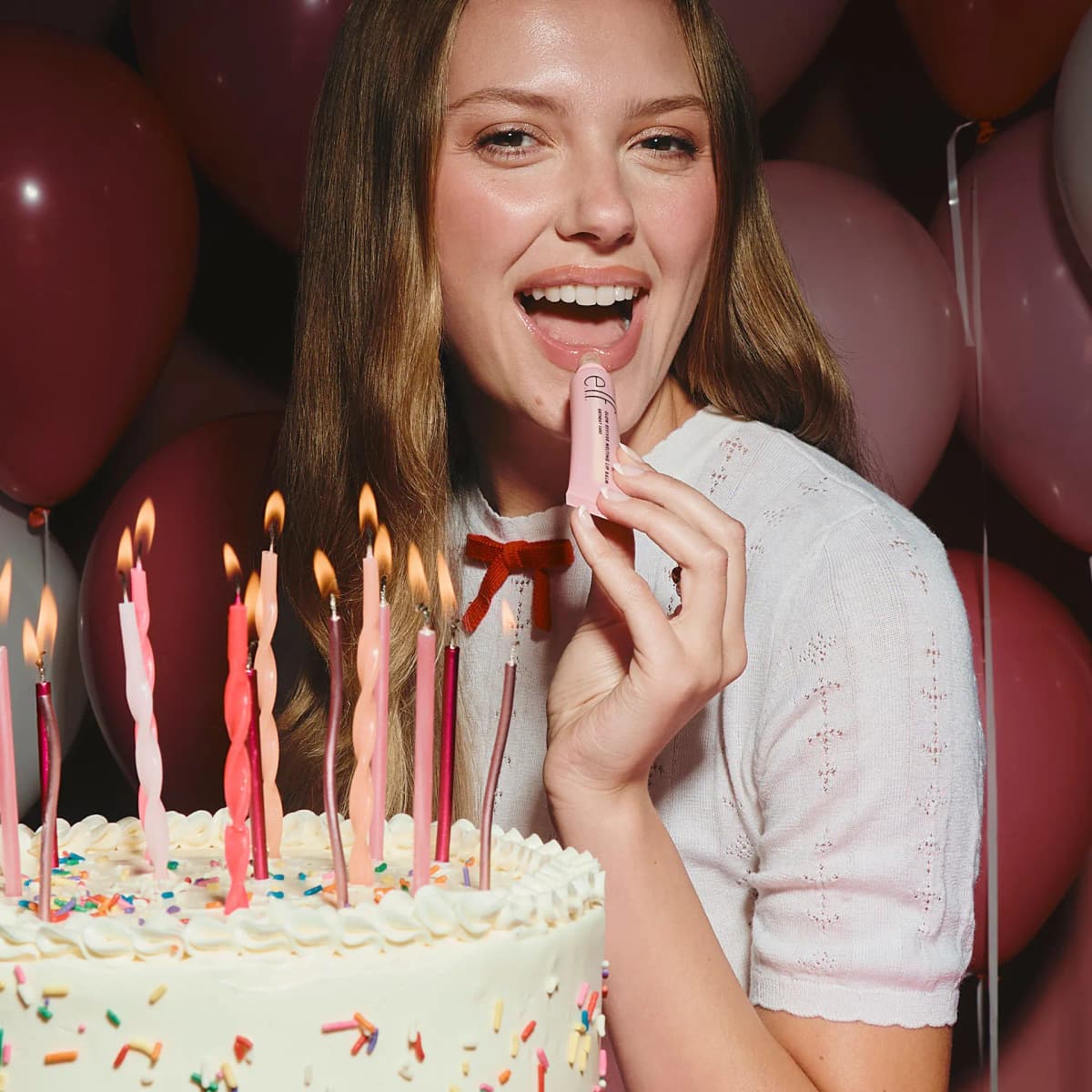 Smiling model applies the pink tube balm beside a candlelit sprinkle cake with burgundy and pink balloons