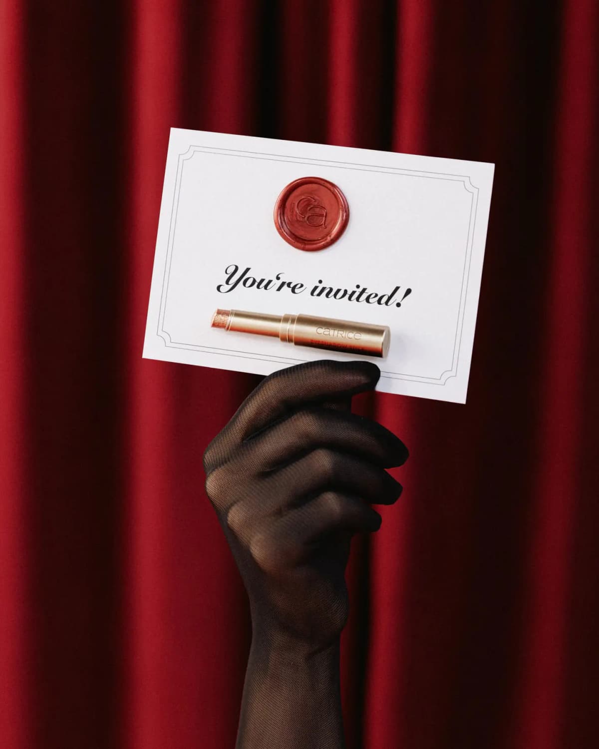 Black-gloved hand holds a wax-sealed You're invited! card with a gold Catrice eyeshadow stick against red curtains