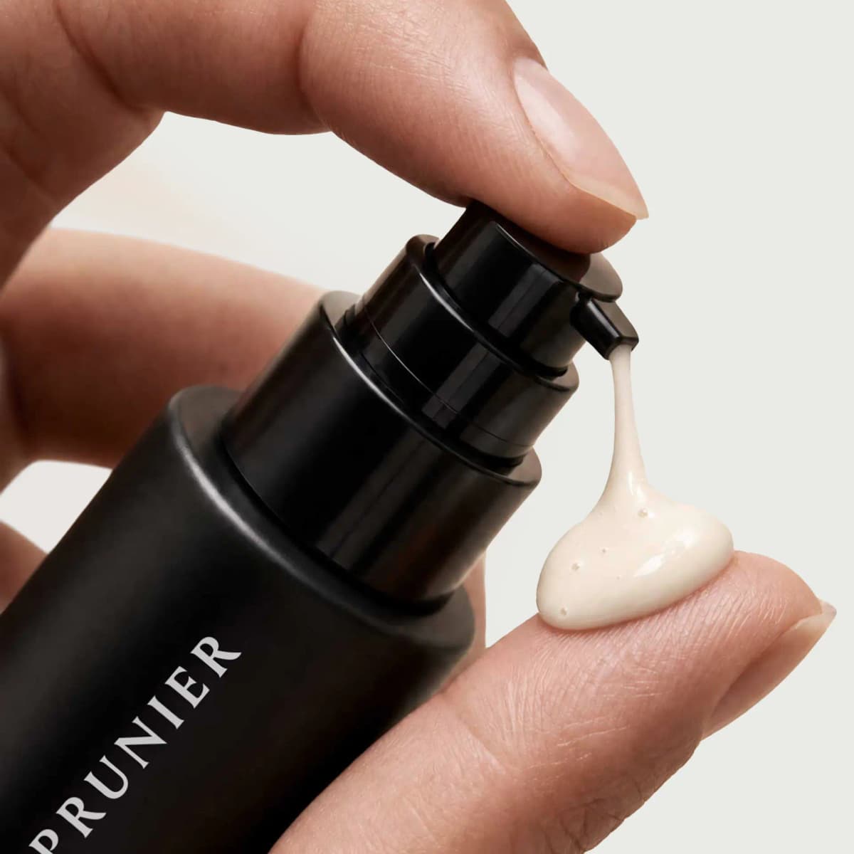 Hand pressing the black pump to dispense a creamy ivory dollop onto a fingertip