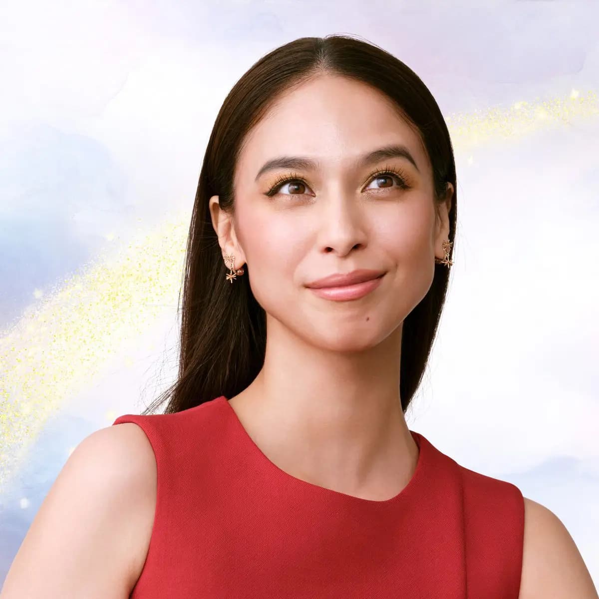Smiling model with sleek dark hair, golden eyeshadow and red sleeveless top against a pastel sky with a gold glitter trail