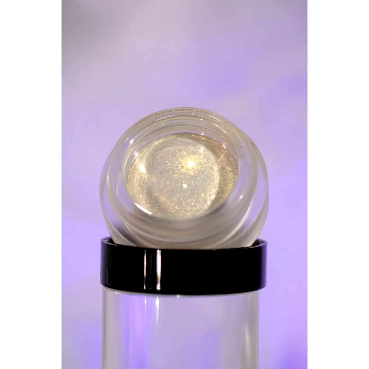 Open glass jar of shimmering champagne-gold cream product resting on a clear acrylic stand against a lilac backdrop