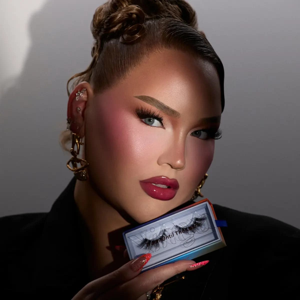 NikkieTutorials in a black blazer with gold earrings, holding a Nimya lash box near her face under dramatic lighting.