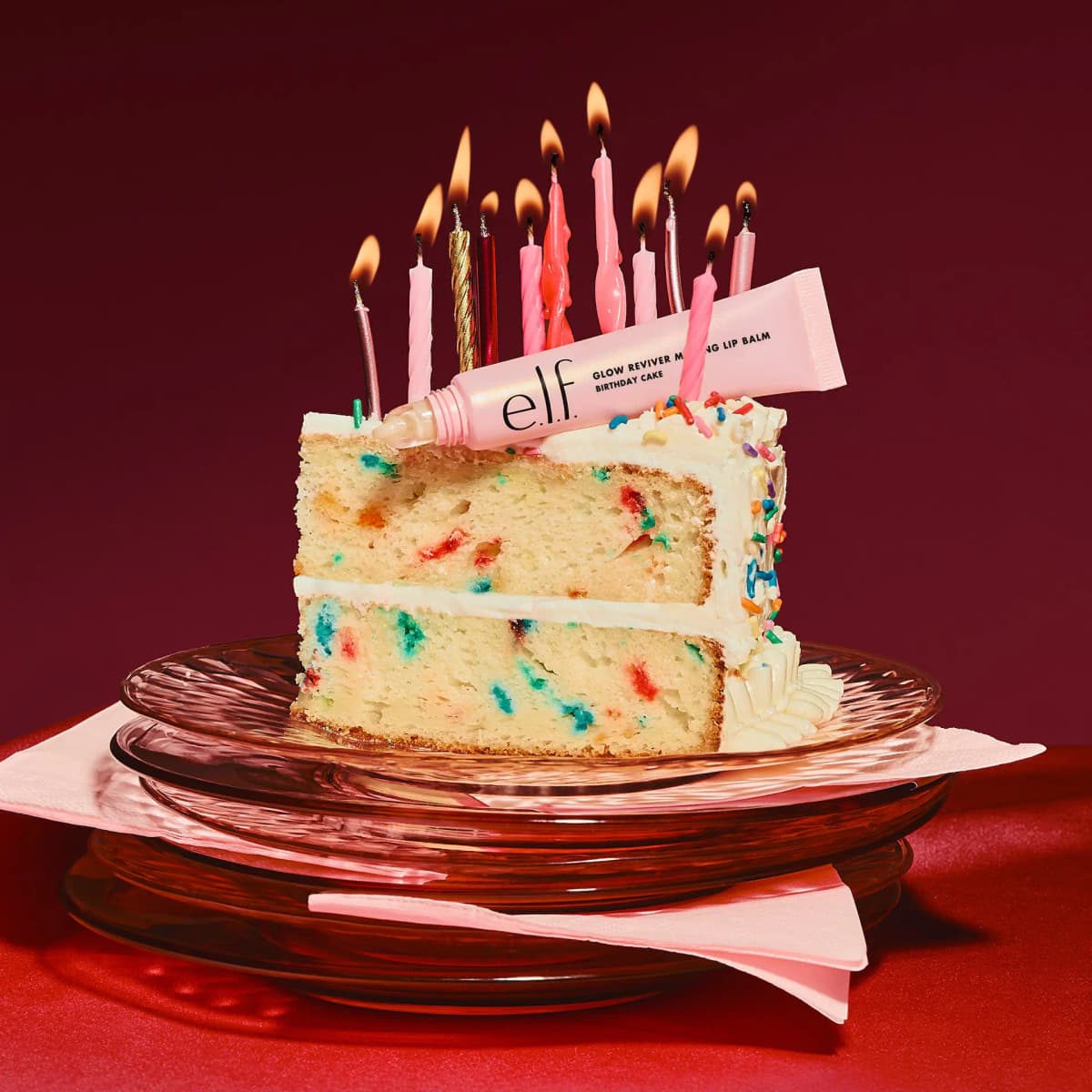 Pink lip balm tube on a funfetti cake slice with lit pink and gold candles on a dark red backdrop