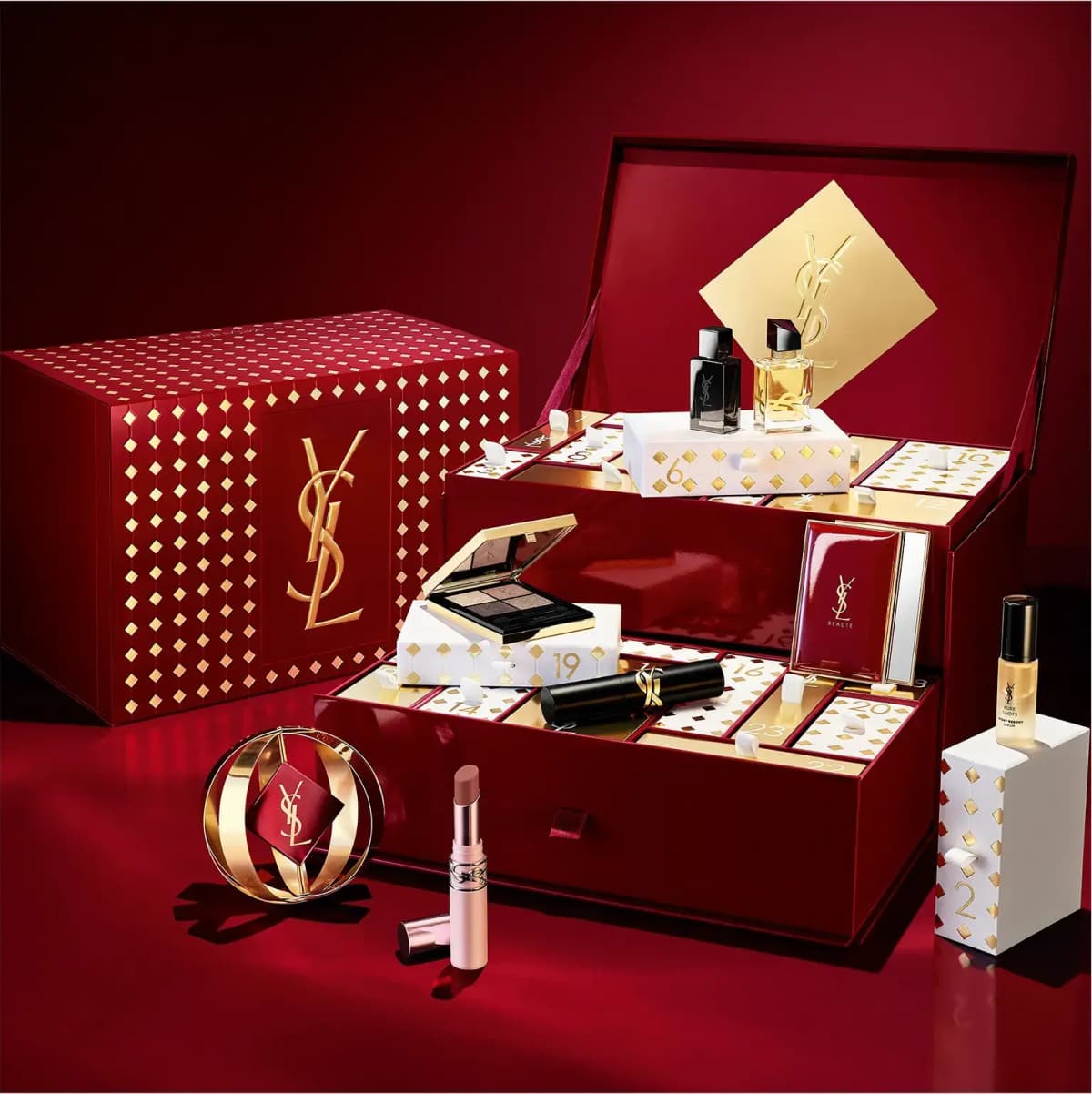 YSL Advent Calendar open on a red backdrop, showing fragrance minis, an eyeshadow quad, lipstick, serum and a gold bauble.