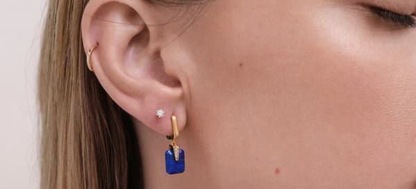Model's ear wearing a tiny stud, a small gold hoop and a hoop with a blue lapis charm
