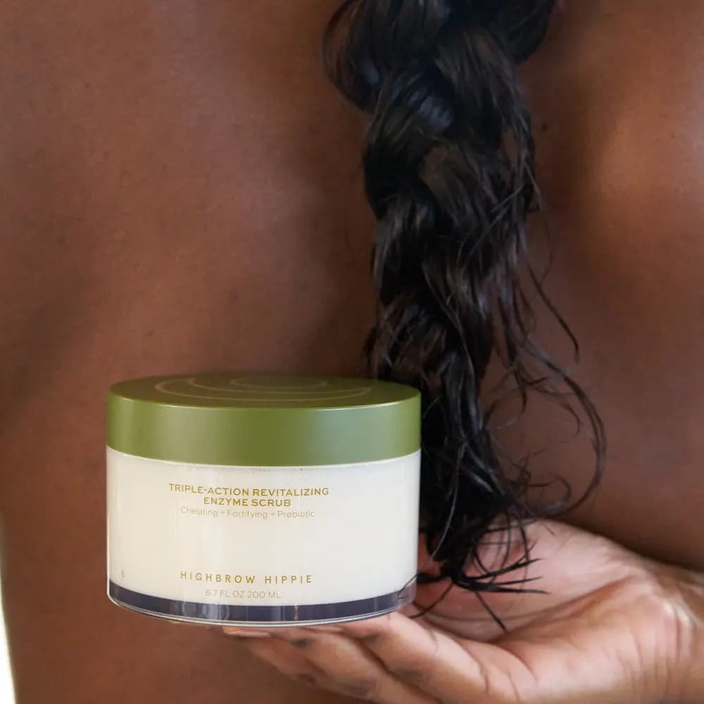 Hand holding the lidded jar of enzyme scrub in front of bare dark skin and a damp twisted dark braid