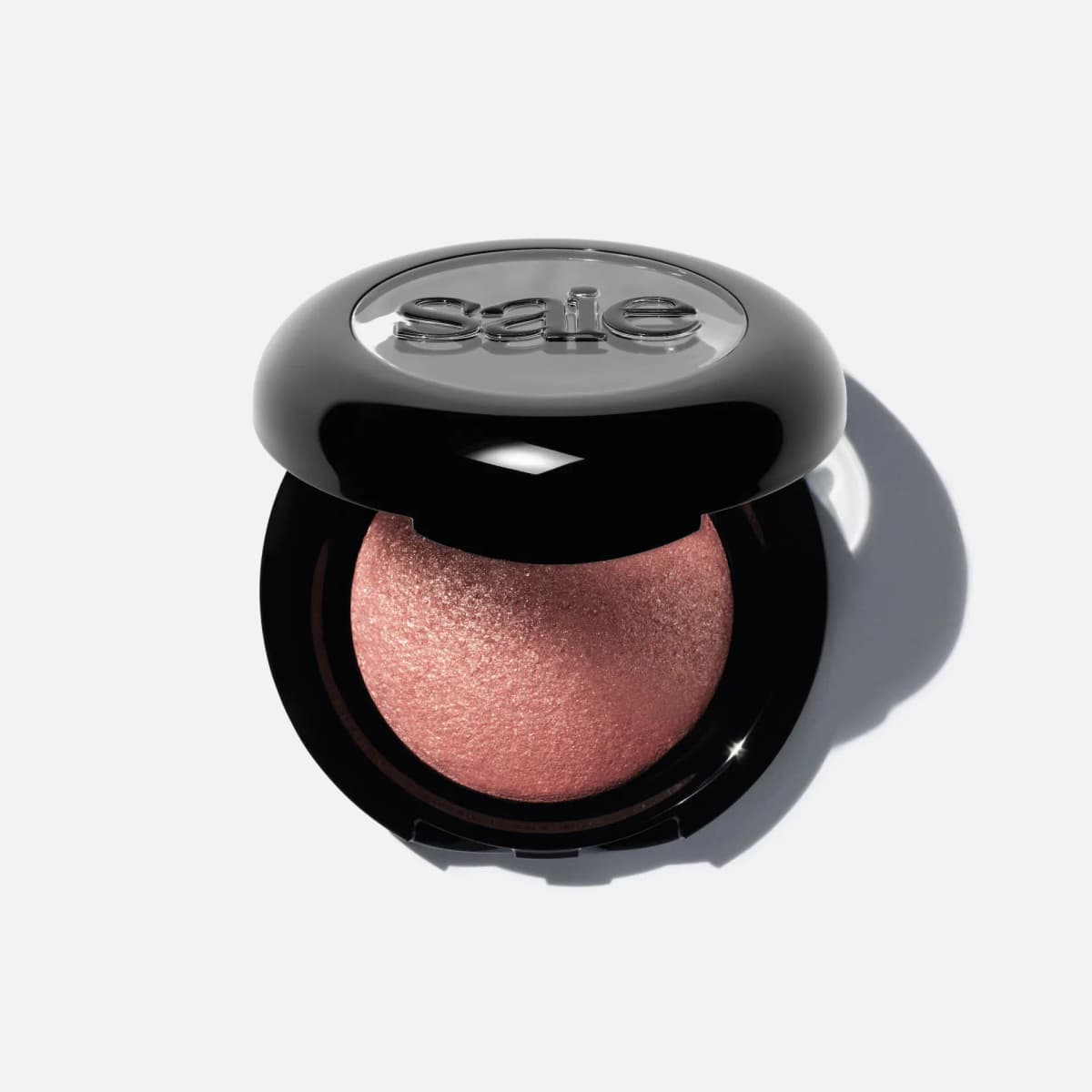Open black Saie compact showing a domed, shimmering rose-pink baked eyeshadow with clear logo lid