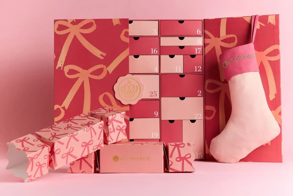 GLOSSYBOX Christmas Collection 2026 Advent Calendar opened to numbered drawers, beside pink crackers and a pink stocking