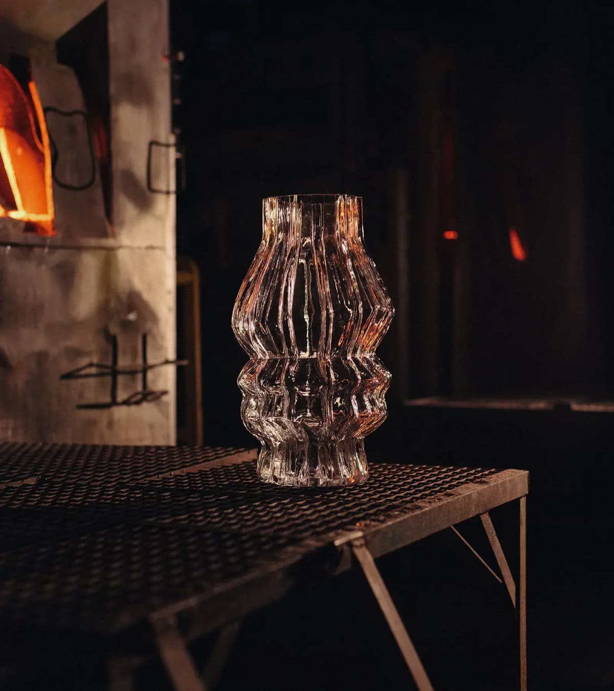 Tall wavy clear glass hurricane on a metal grate table beside a glowing glassworks furnace