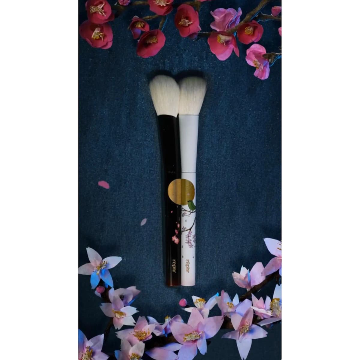 Black and white brushes side by side forming a gold moon with green bird motif, framed by paper plum and cherry blossoms