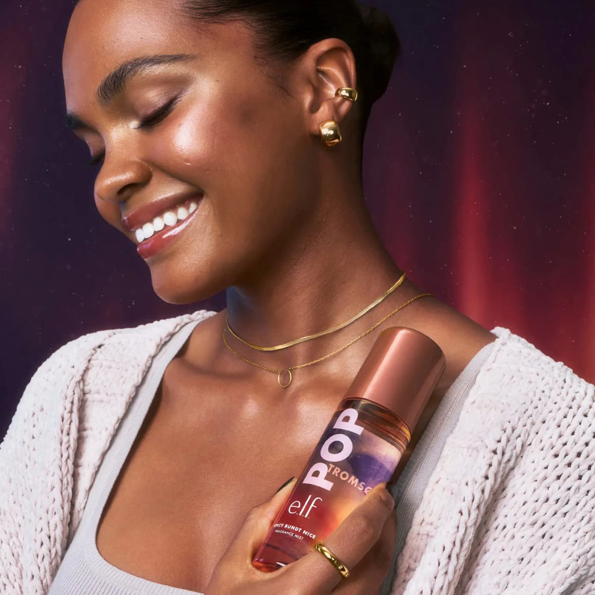 Smiling model in a cream knit cardigan and gold jewellery hugs the e.l.f. POP Spicy Bundt Nice mist to the chest.