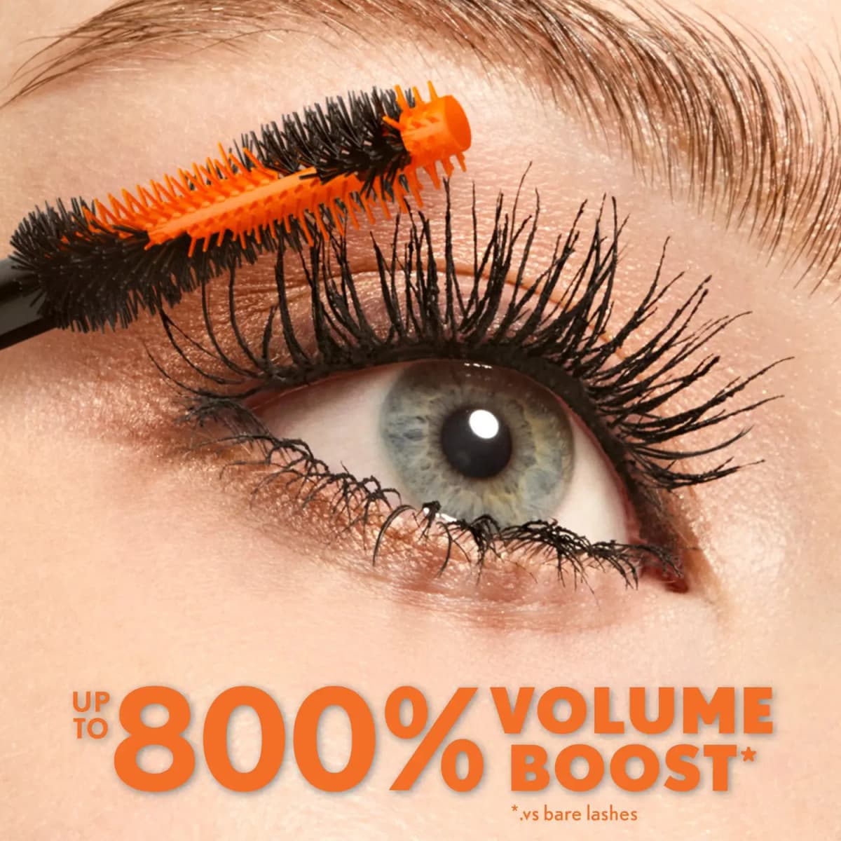 Orange and black brush coating voluminous black upper lashes on a grey-blue eye, claiming up to 800% volume boost