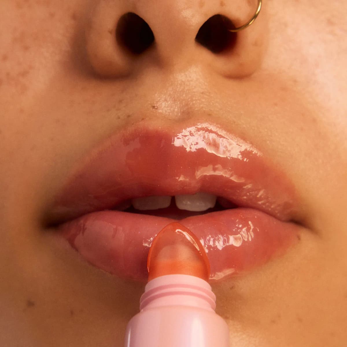 Close-up of glossy lips as a pink tube's peach-tinted tip applies the treatment, with a gold nose ring above.