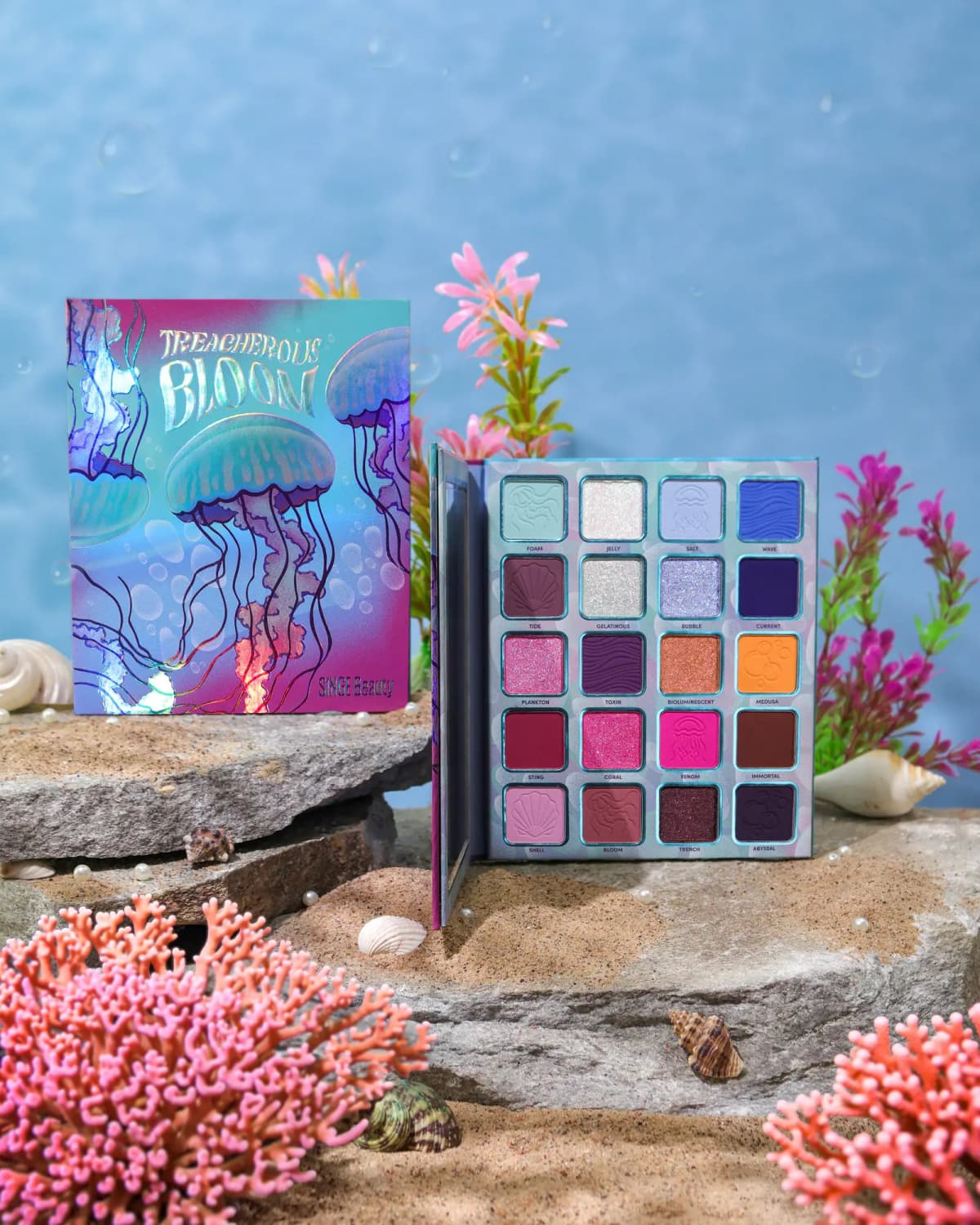 Holographic jellyfish-print Treacherous Bloom palette box beside open 20-pan eyeshadow palette on rocks with pink coral