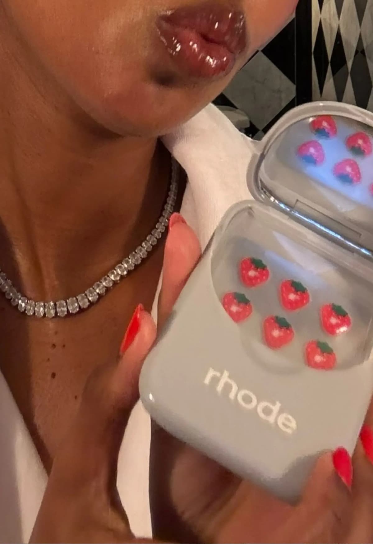 Person with glossy lips and diamond necklace holding the open grey case with strawberry stickers, red nails