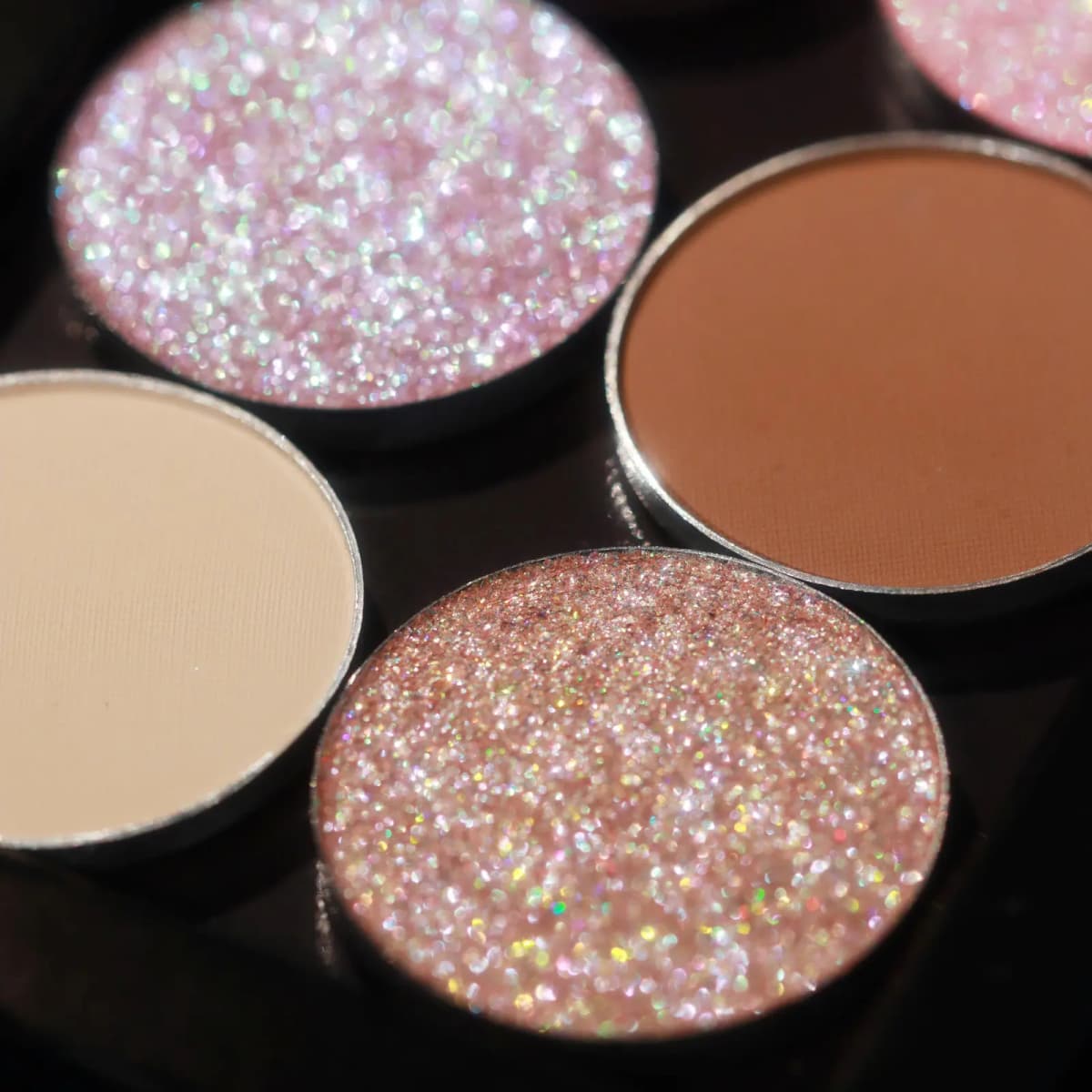 Close-up of four eyeshadow pans showing pink glitter, rose gold sparkle, matte nude, and matte brown finishes
