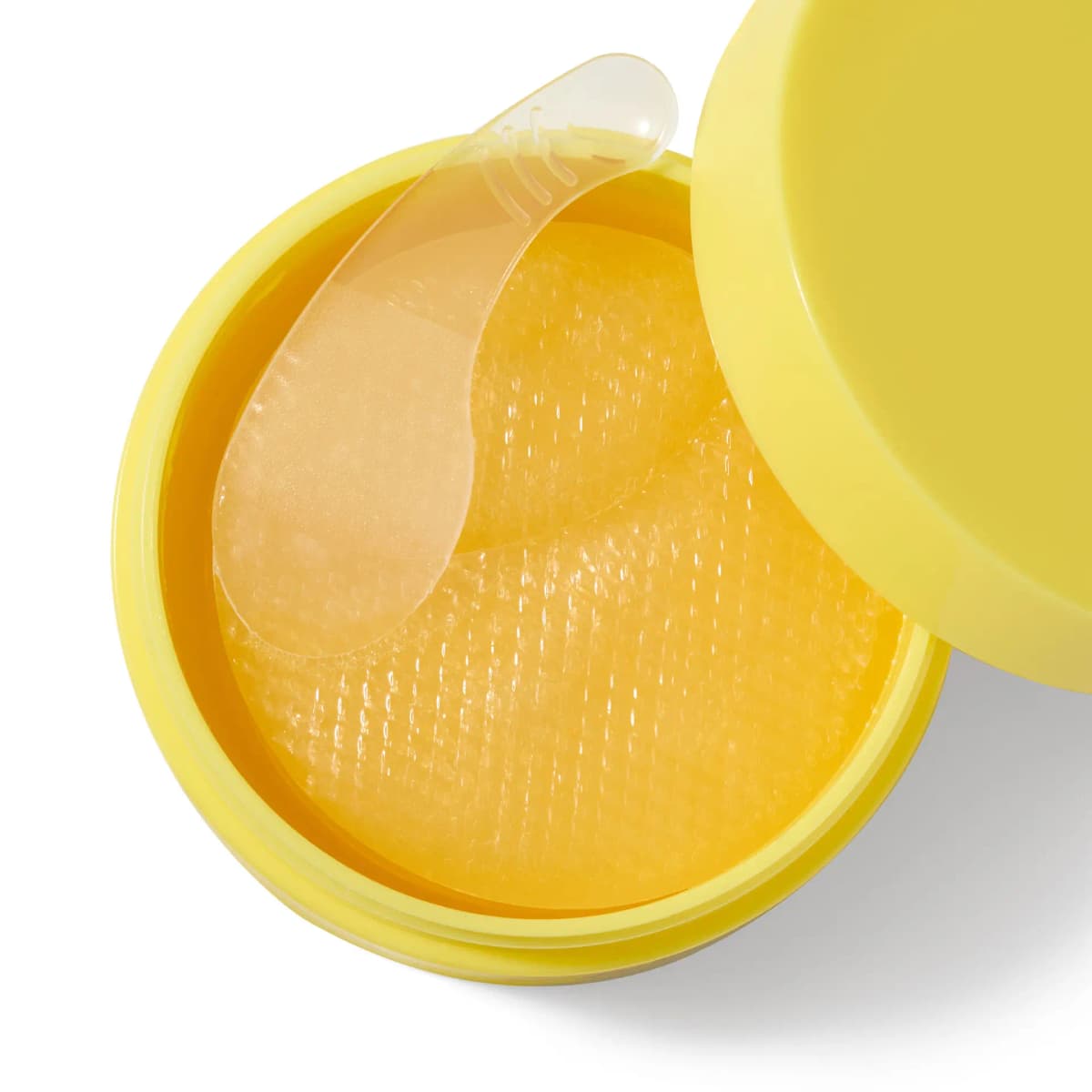 Open yellow jar of stacked golden textured gel patches with a clear spatula lifting one, lid beside it
