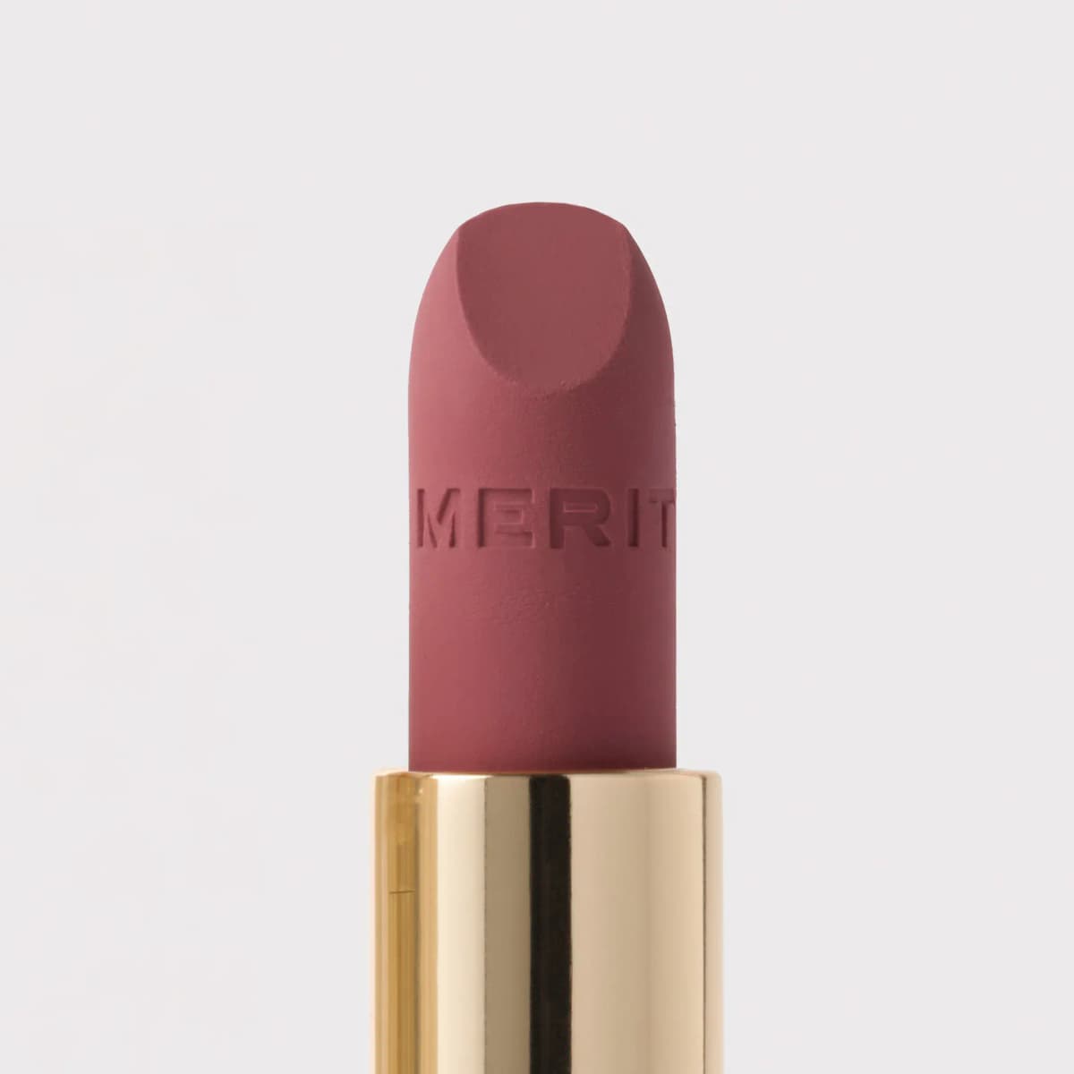 Close-up of a muted rose-mauve matte lipstick bullet embossed with MERIT, rising from a shiny gold tube.
