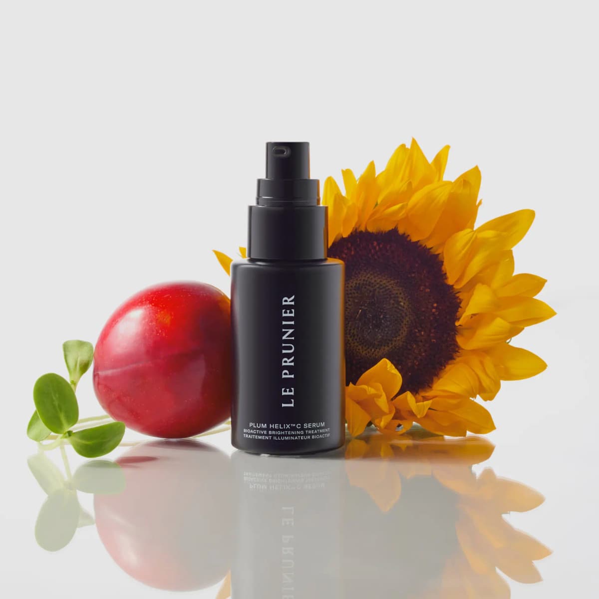 Black serum bottle on a reflective surface between a red plum, a yellow sunflower and green sprouts