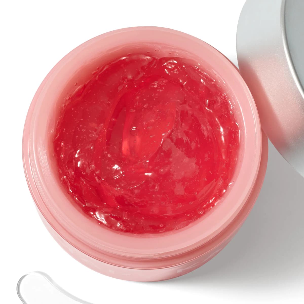 Open pink jar from above showing glossy red gel moisturizer with tiny air bubbles, silver lid and clear spatula nearby