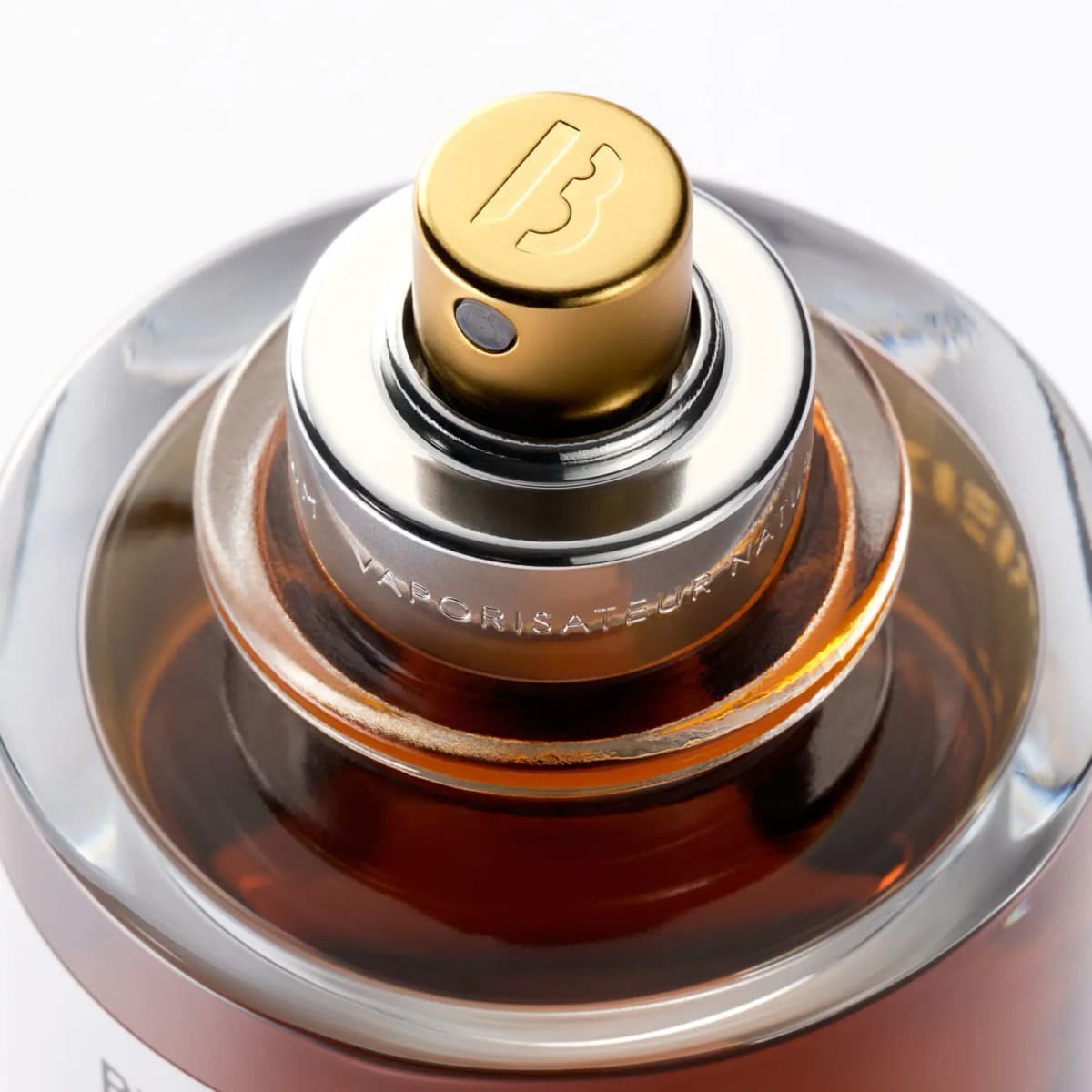 Overhead close-up of the gold B-logo spray nozzle and silver collar engraved Vaporisateur Natural over amber liquid