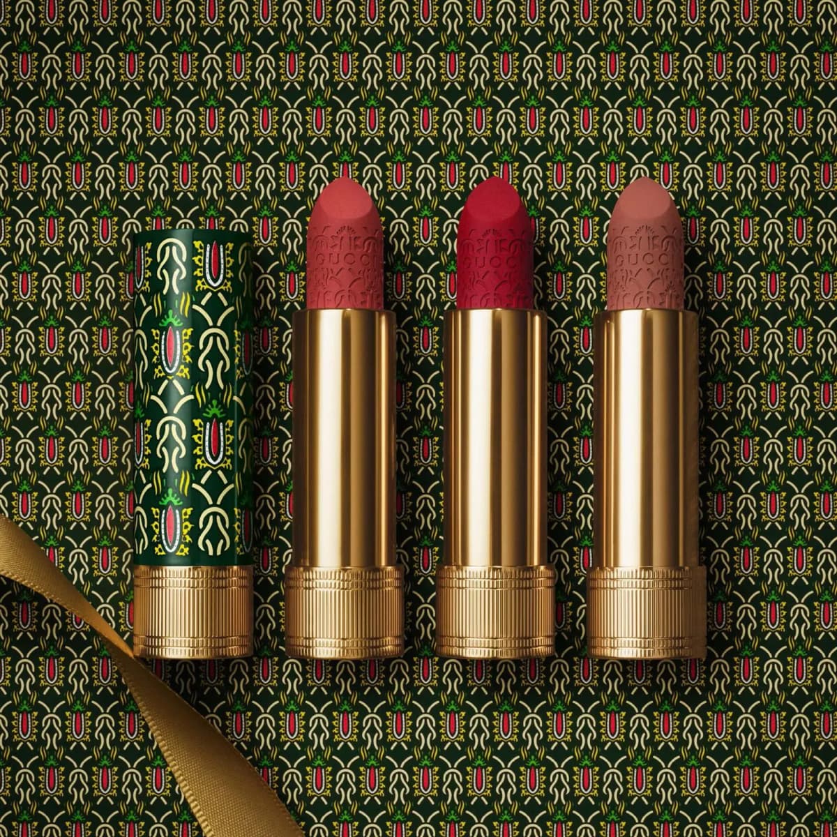 Three engraved matte lipsticks in rose, red and nude in gold tubes beside a patterned green cap on matching wrapping paper