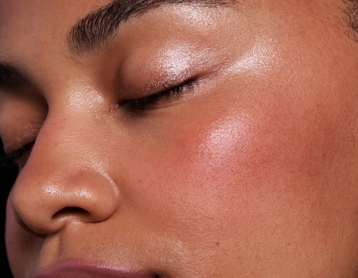 Close-up of a medium-skinned model with closed eyes, a warm pink cheek flush and glossy sheen on lid and cheekbone