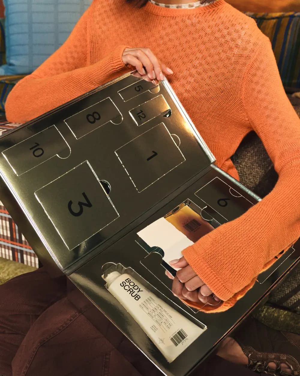 Model in orange knit sweater holding the open mirrored calendar, body scrub tube in one compartment