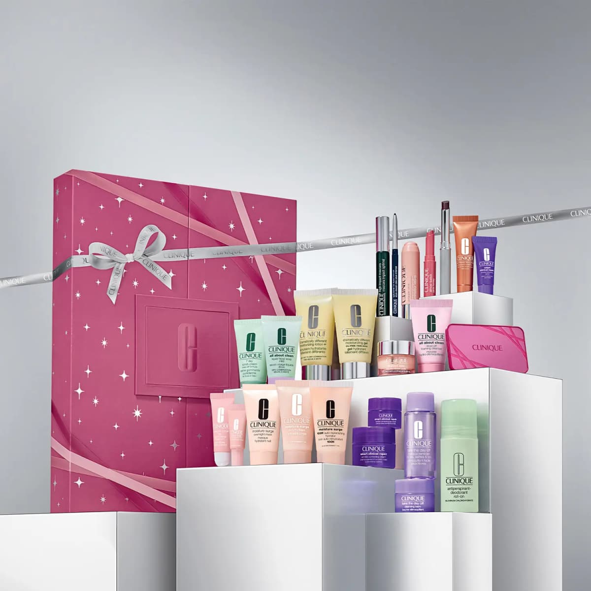Pink Clinique calendar box beside its products arranged on white plinths, with Clinique-branded silver ribbon behind