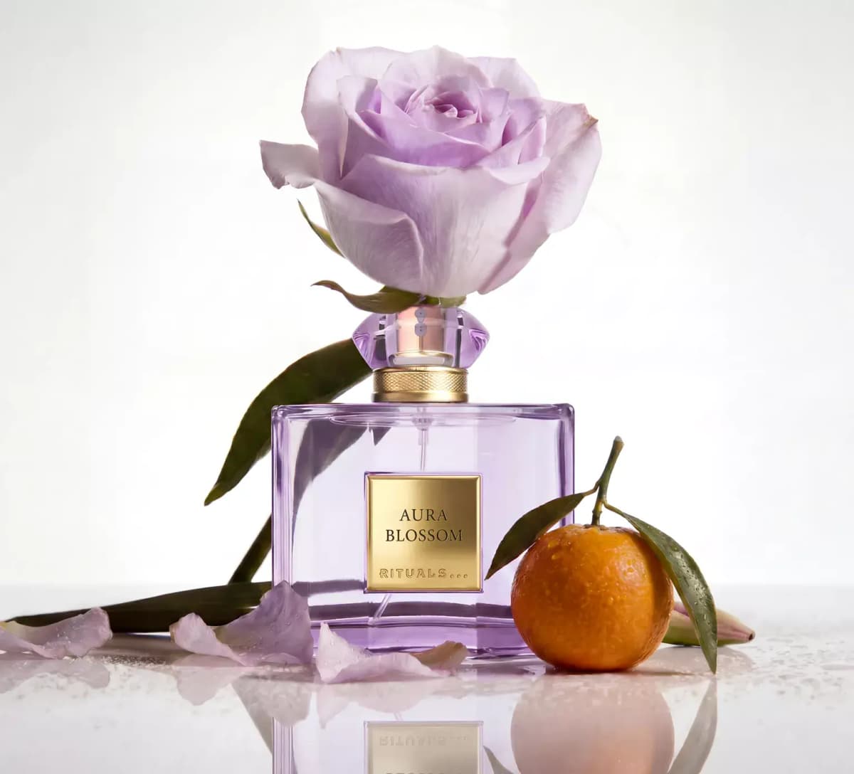 Lilac square Rituals Aura Blossom bottle with gold label, a lavender rose on top, a mandarin and fallen petals