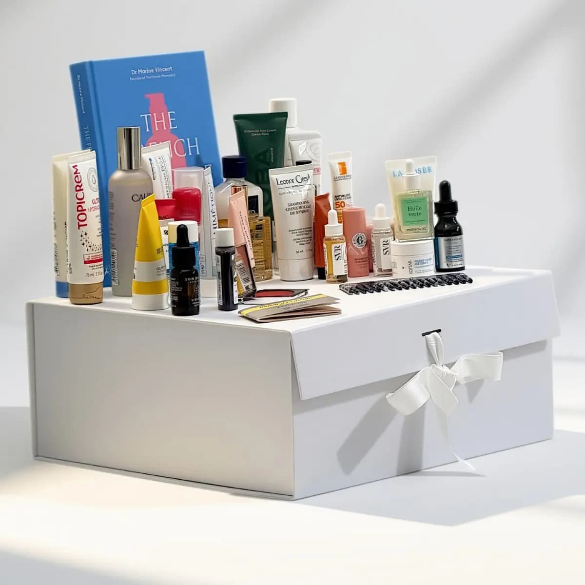 White magnetic keepsake box with white ribbon bow, topped with an array of French pharmacy skincare products and a blue book