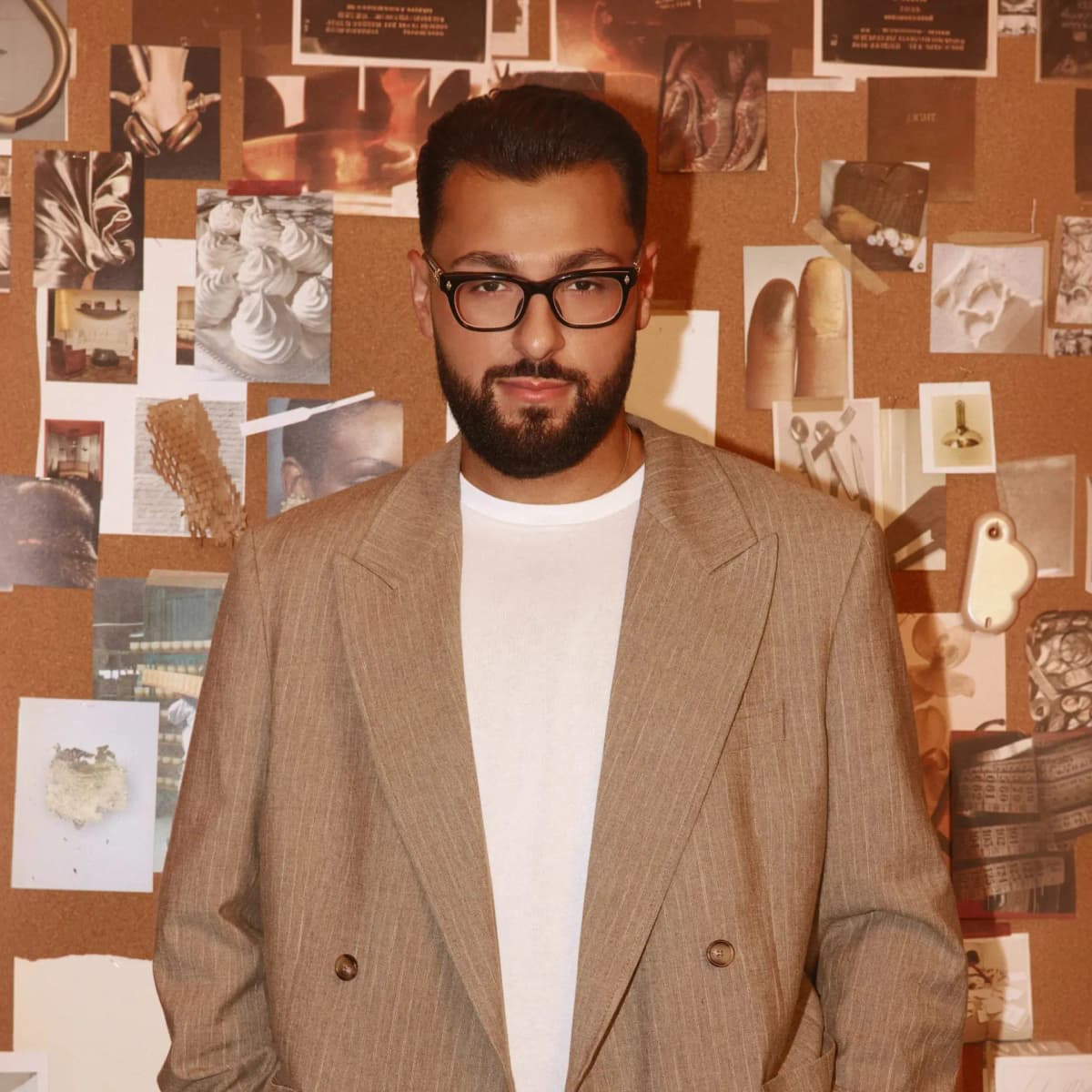 Paul Reacts in glasses and a beige double-breasted blazer before a corkboard mood board of textures and photos