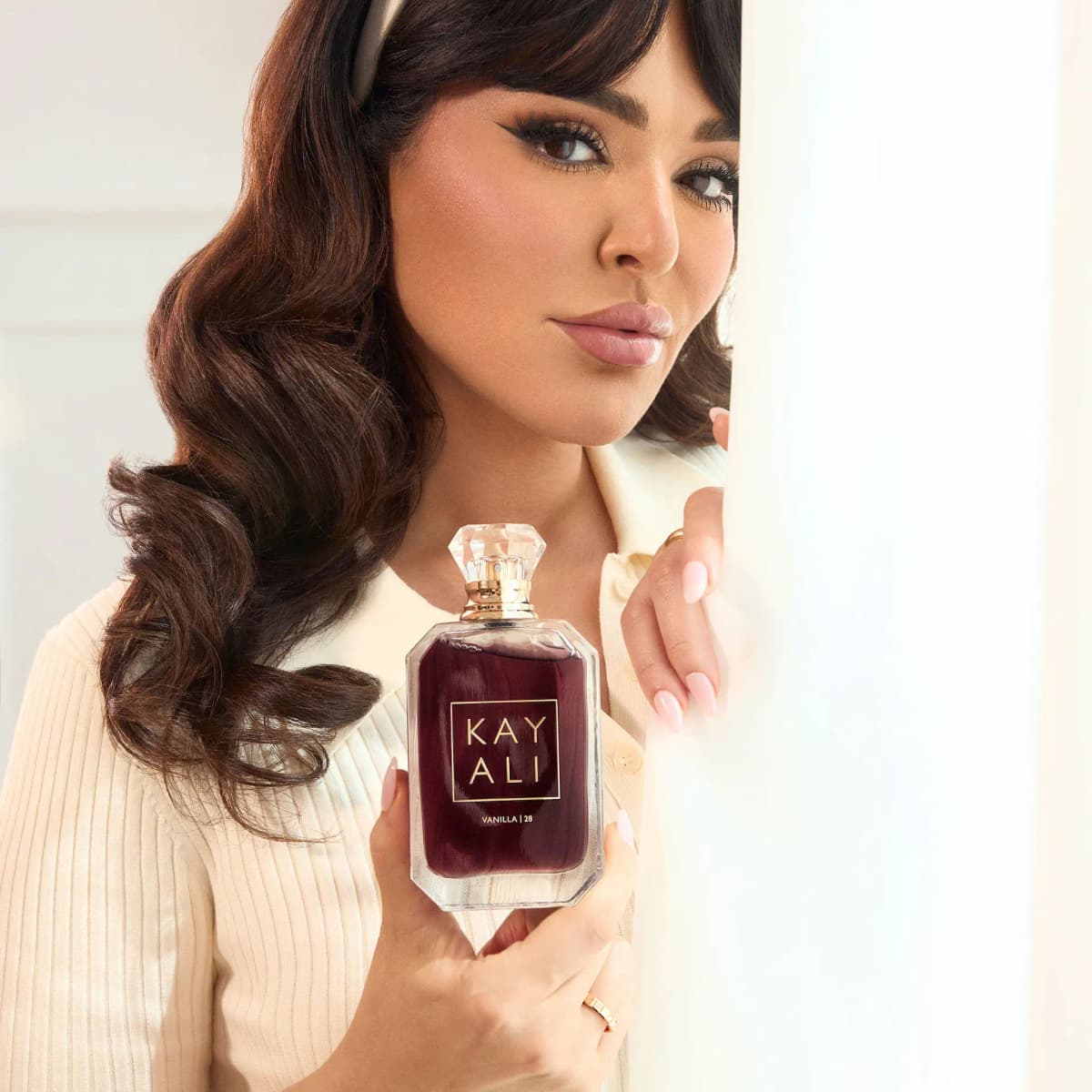 Woman with wavy brown hair in a cream cardigan holding the Vanilla | 28 bottle up beside her face