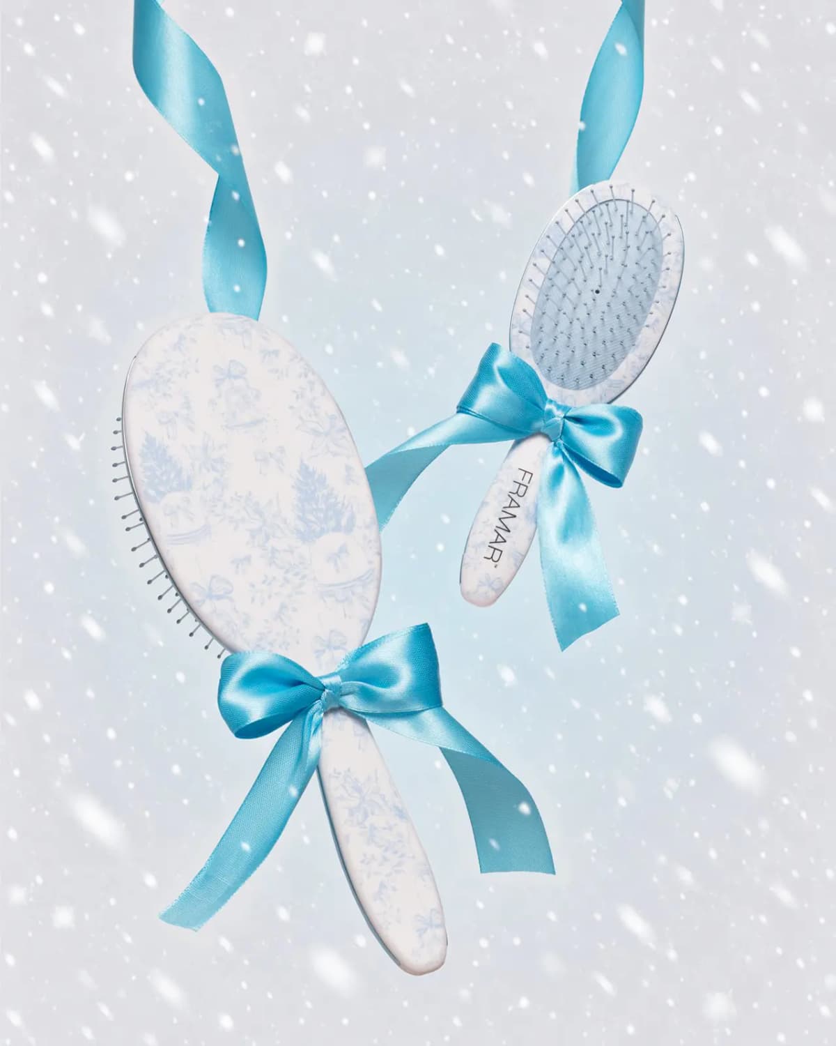 Two FRAMAR toile brushes tied with glossy turquoise satin ribbon bows against a snowy pale-blue background