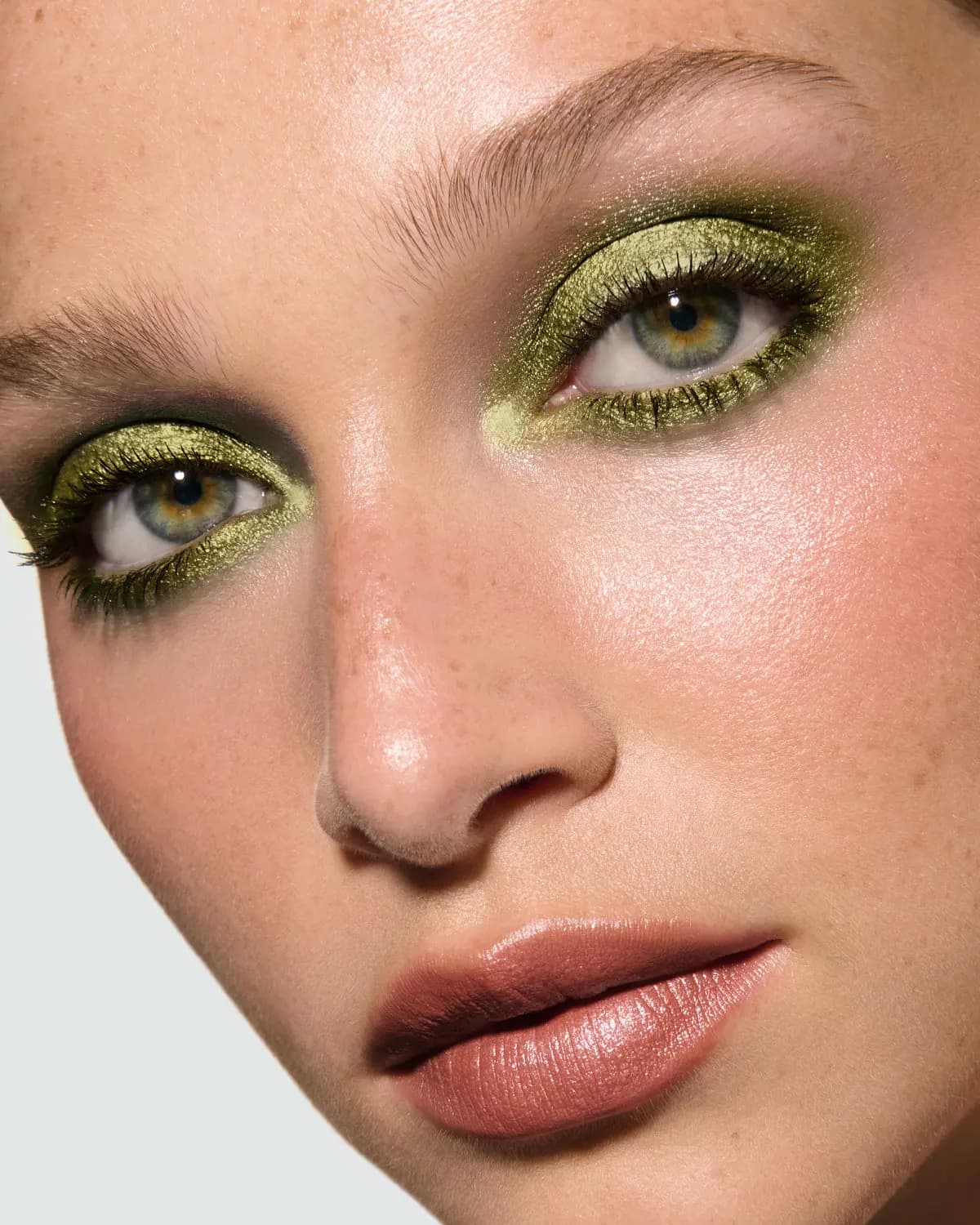 Close-up of a freckled model wearing metallic olive green eyeshadow, rosy cheeks and a nude pink lip