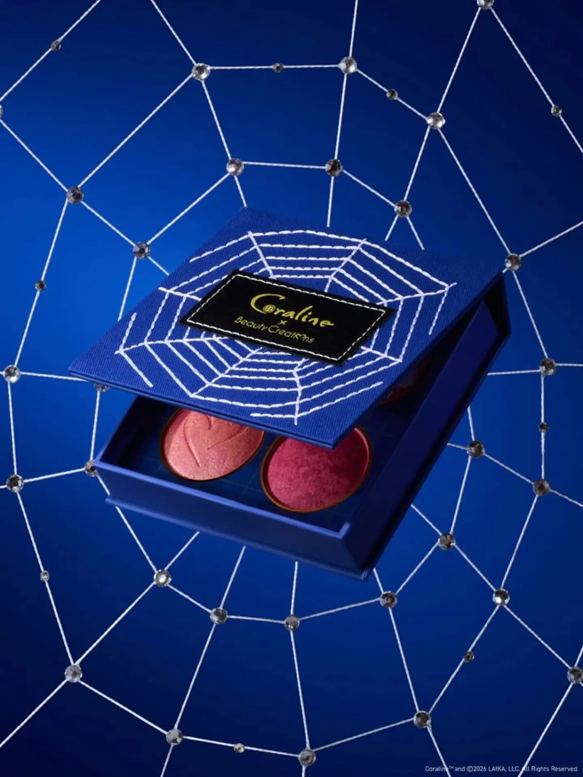Blue spiderweb-stitched face palette open to reveal two shimmery pink and berry powders, set against a jeweled web backdrop