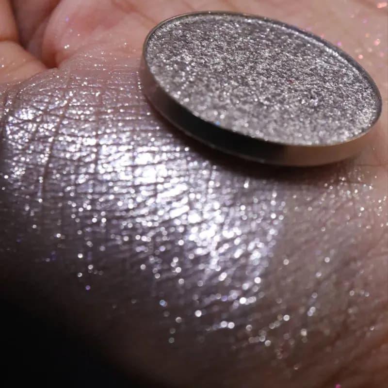 Close-up of a silver metallic shimmer shadow pan beside a glittering silver swatch with a lilac sheen on a hand.