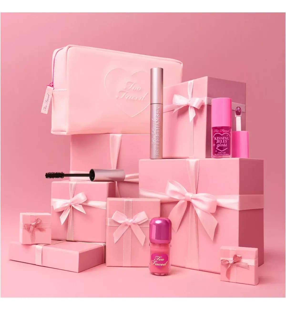 Pink pouch, mascara, lip gloss and liquid blush styled among pink gift boxes tied with satin ribbon bows