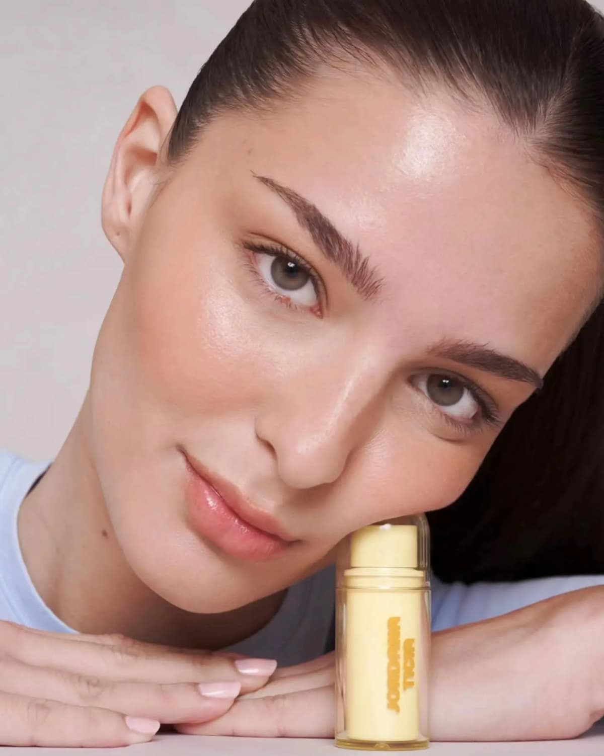 Brunette model with slicked-back hair rests her cheek on a capped yellow stick, skin dewy and bronzed