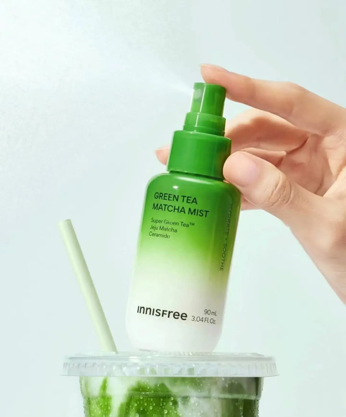Hand spraying the green-to-white innisfree Green Tea Matcha Mist bottle above an iced matcha latte with a pale green straw