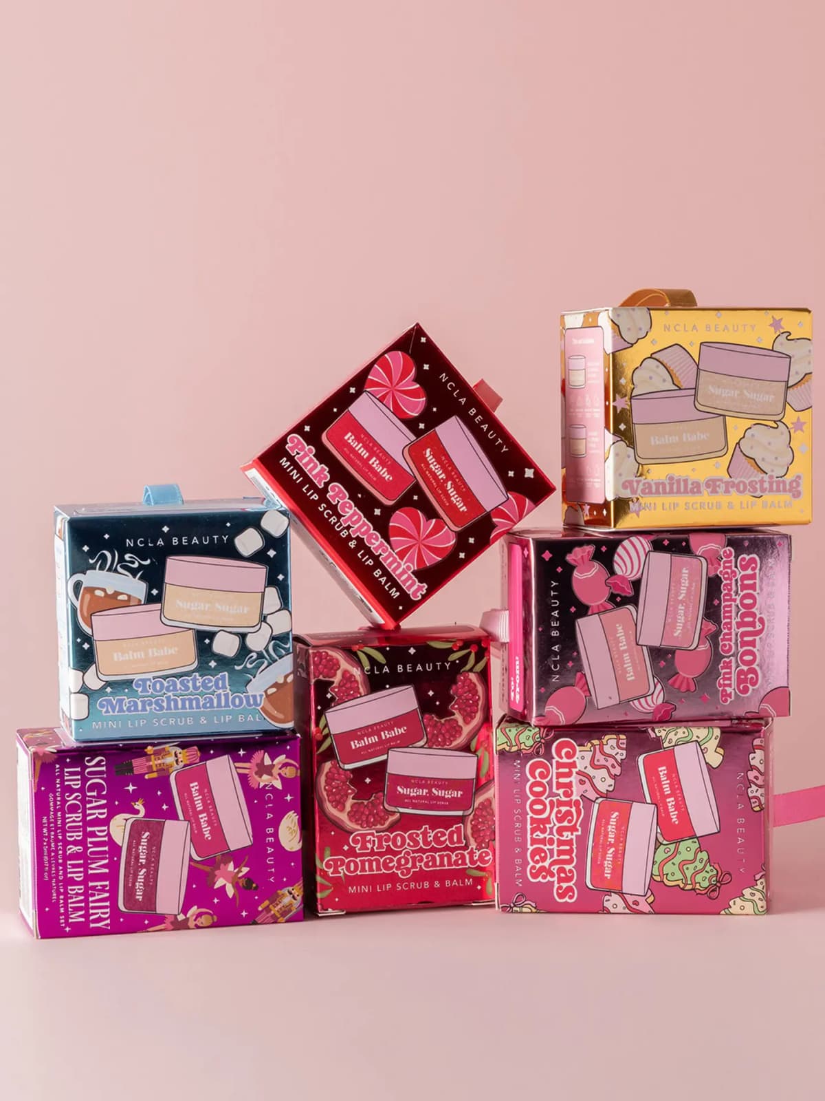 NCLA Beauty Holiday 2026 Lip Scrub & Balm Ornaments: seven foil gift boxes stacked on a pink backdrop